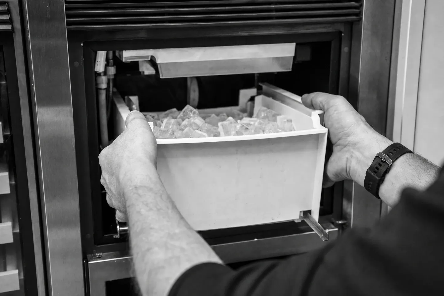 Black-and-white view of hands lifting an ice bin full of cubes out of the ice-maker compartment of a built-in unit