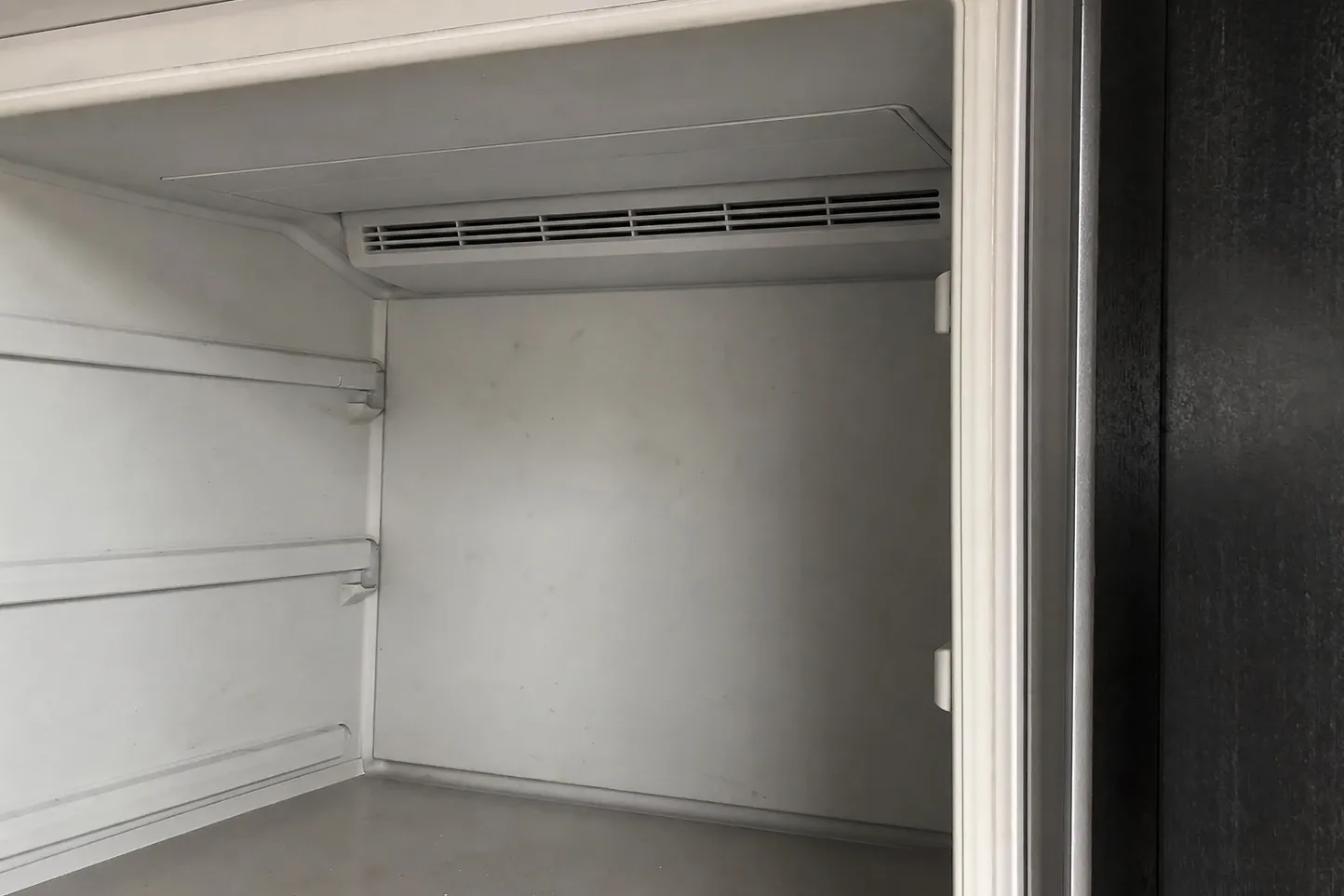 The inside of an emptied white compartment: a louvred air vent set under the top shelf, moulded shelf rails down the left wall and the edge of the door seal at the right. Labelled on the photograph: door seal edge, shelf rails, air vent.