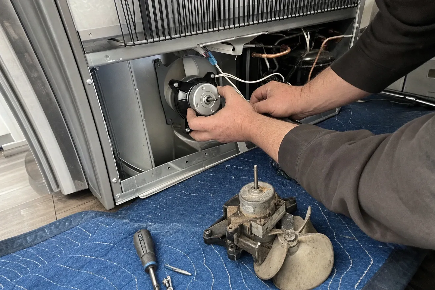 Hands seating a clean fan motor into its bracket in an open bay, condenser fins across the top of the opening and a compressor to the right, the dust-caked old motor with its blade and a screwdriver on a blue quilted blanket below. Labelled on the photograph: new motor in its bracket, condenser fins, compressor.