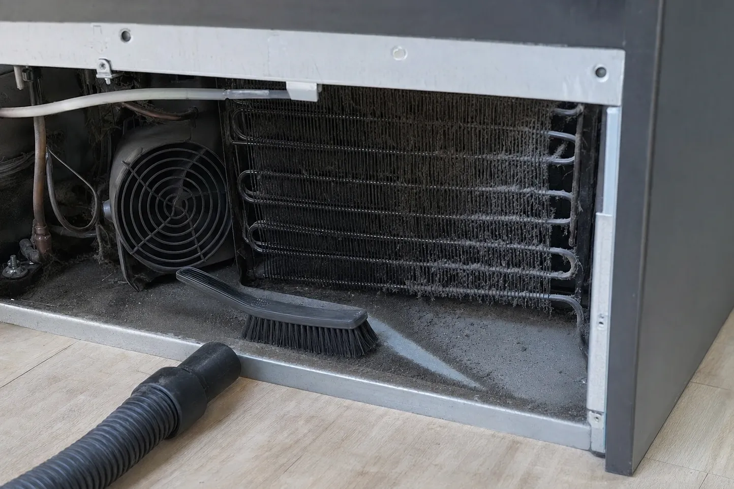 A condenser coil furred grey with lint behind the removed lower grille of a dark cabinet, the condenser fan in its housing to the left and a brush and vacuum hose waiting on the floor. Labelled on the photograph: lint on the base pan, condenser coil under lint, condenser fan.
