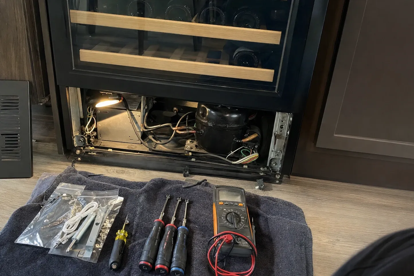 A glass-door wine cabinet with its lower access panel removed, a work light shining on the compressor and copper lines inside the bay, and a bagged parts kit, screwdriver, three nut drivers and a multimeter laid out on a towel below. Labelled on the photograph: compressor, work light, parts kit, still bagged.