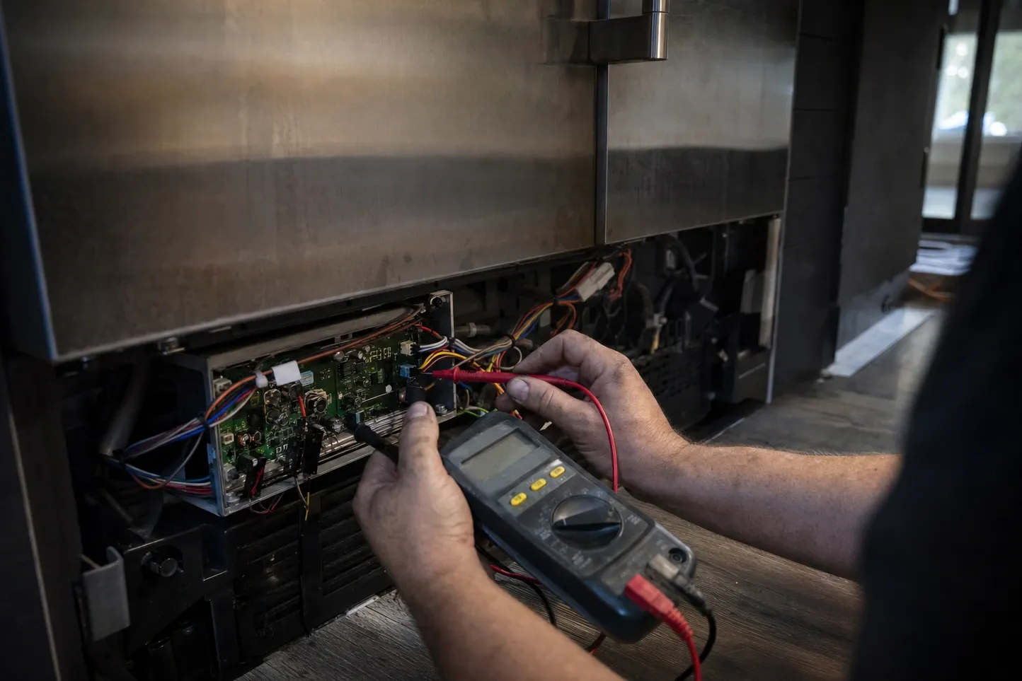Hands holding a black multimeter and a red probe against a green circuit board in the open lower bay of a stainless cabinet, coloured wiring running away to the right. Labelled on the photograph: circuit board, red probe, multimeter.